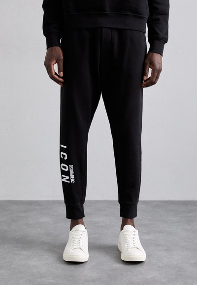 Pantalon de survêtement noir avec des jambes fuselées et des poignets côtelés. Présente un logo blanc sur la jambe gauche. Associé à des baskets blanches.