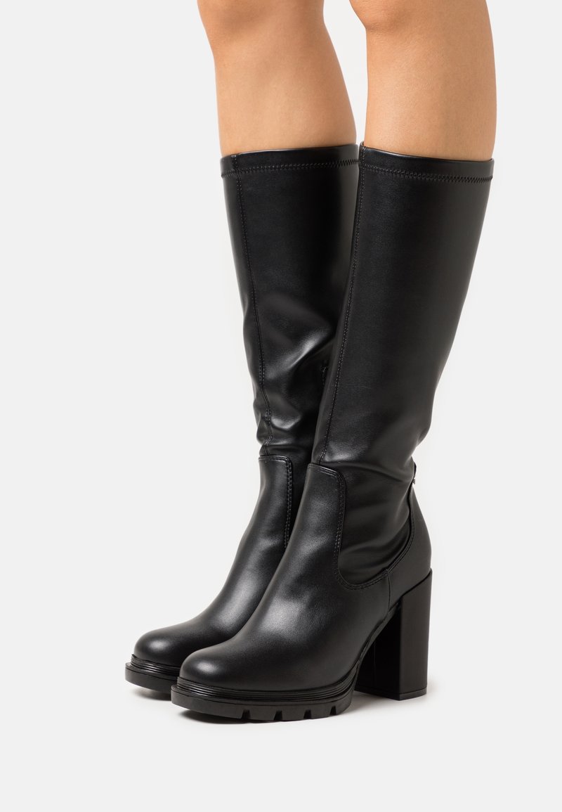 Bottes en cuir noires montant jusqu'aux genoux avec talons carrés épais et semelles à crampons, portées sur des jambes nues sur un fond blanc uni.