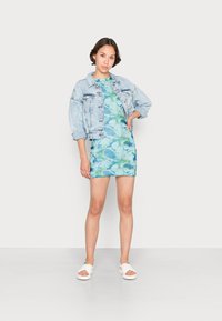 Casaco de ganga azul-claro sobre um vestido mármore em tom teal. Modelo a usar sandálias brancas, de pé sobre um fundo branco, com as mãos na anca.