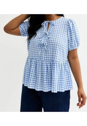 Kobieta ubrana w niebiesko-białą bluzkę w kratkę z krótkim rękawem, trzema kokardkami z przodu i falbaną u dołu, w połączeniu z ciemnoniebieskimi dżinsami.