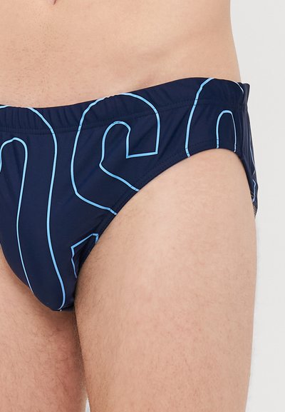 Nahaufnahme einer Person in navyblauen Schwimmslips mit hellblauen umrandeten Buchstaben auf dem Stoff, die den Hüft- und Oberschenkelbereich zeigt.