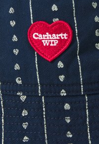 Țesătură albastră cu modele în formă de inimi albe și linii verticale. Are o aplicație în formă de inimă roșie cu textul "Carhartt WIP" în alb.