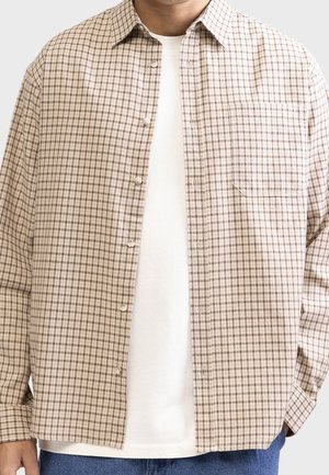 Man wearing beige and brown checkered button-up shirt over white T-shirt and blue jeans, standing against light background.