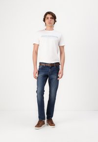 Jeans de mezclilla en azul oscuro con un corte ajustado, combinados con una camiseta de algodón blanca que presenta texto azul. Zapatillas de cuero marrón y un cinturón marrón completan el look.