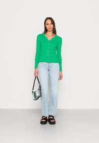 Cardigã verde com nervuras, com uma barra rendada e botões, combinado com jeans de perna reta azul claro e uma pequena bolsa preta e branca.