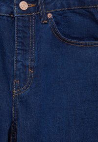 Los vaqueros de mezclilla azul oscuro presentan costuras en contraste de color naranja, un botón de metal y un bolsillo frontal con una apertura curva.
