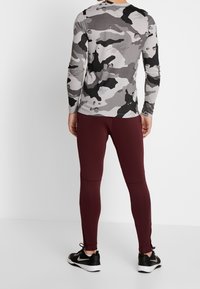Langarmige grijze camouflage shirt van textuurstof, gecombineerd met strakke bordeaux leggings met details op de knie en zwarte schoeisel.