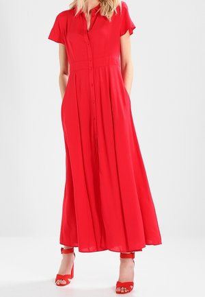 Žena v lehké červené maxi šatech s krátkými rukávy a červených sandálech na podpatku, stojící před jednobarevným pozadím.