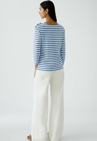 Blau-weißer gestreifte Langarmoberteil mit einem Bootsausschnitt und Schulterknopfdetails, kombiniert mit lockeren weißen Hosen.