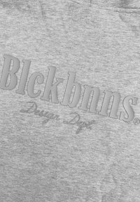 Grijze sweatshirt met verhoogde, gedempte tekst die "Blckbnns" en "Design Dept" in contrasterende kleuren leest, met een gladde stoftextuur.
