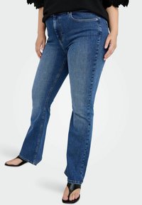 Denim bikses vidēji zilā krāsā ar izplestām kājām, piecām kabatām un ar rāvējslēdzēju. Audumam ir nedaudz teksturēts virsma.