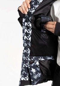 Schwarz-weiße Jacke mit geometrischen Mustern, ausgestattet mit Netz- und Stofftaschen. Das Material wirkt leicht und atmungsaktiv. Innenfutter sichtbar.