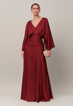 Vrouw met een lange bordeauxrode jurk met brede mouwen en geplooide taille, met in haar hand een kleine gouden clutch tegen een effen lichte achtergrond.