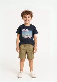 Marineblauwe katoenen t-shirt met een grafische print, gecombineerd met khaki shorts met cargovakken, en witte sneakers. Kind met een glimlach.