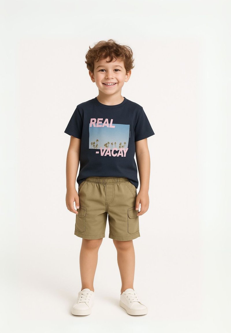 Marineblauwe katoenen t-shirt met een grafische print, gecombineerd met khaki shorts met cargovakken, en witte sneakers. Kind met een glimlach.