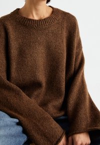 Pull en tricot marron avec un col rond côtelé, des manches larges et une texture douce. Associé à un jean en denim bleu, mettant l'accent sur un design confortable.