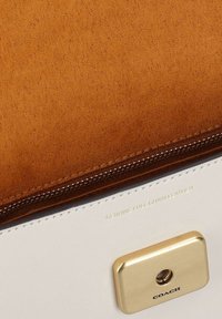 Rabats en daim marron sur sac en cuir crème avec une boucle rectangulaire dorée portant les inscriptions "COACH" et "GENUINE FULL GRAIN LEATHER" embossées au-dessus.