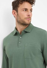 Langärmliges Polohemd aus grünem Stoff, mit Knopfleiste und einer Brusttasche mit kontrastierender Naht. Glatte Textur.