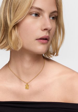 Goldkette mit einem blumenförmigen Anhänger, der ein strukturiertes Design auf einer dünnen Kette aufweist, getragen von einem Modell vor einem neutralen Hintergrund.