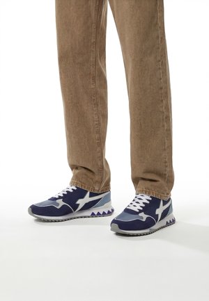 Personne portant un pantalon beige et des baskets bleues, blanches et grises avec une semelle à motif en zigzag, debout sur une surface blanche.