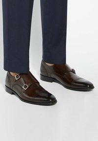 Brown leather double monk strap shoes with silver buckles, featuring a polished finish and a slightly pointed toe design, paired with navy trousers.