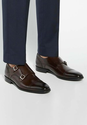 Brown leather double monk strap shoes with silver buckles, featuring a polished finish and a slightly pointed toe design, paired with navy trousers.