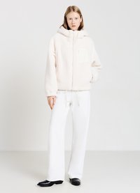 Felpa con cappuccio in morbido tessuto crema, con tasca frontale, orlo elasticizzato e lacci. Abbinata a pantaloni bianchi a gamba larga e stivali neri.