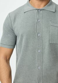 Polo gris à manches courtes présentant une texture douce, un devant à boutons et une poche sur le côté gauche de la poitrine, avec un design de col classique.