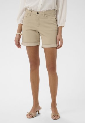 Kvinde iført beige shorts med opsmøgede kanter, en hvid bluse med lange ærmer, guldbøjle og beige åbne hæle stående mod en ensfarvet baggrund.