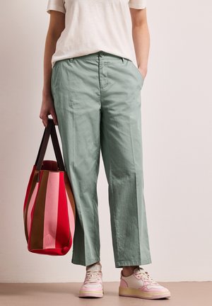 Persona con pantalones verde claro, camiseta blanca de manga corta, zapatillas rosa y blancas, sosteniendo un bolso de mano a rayas rojas y marrones.