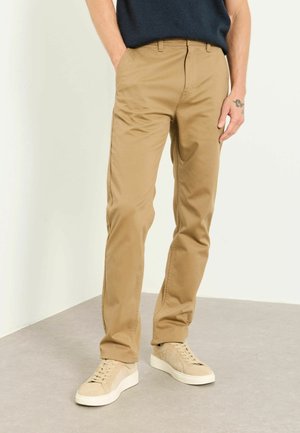Mann, der beige gerade geschnittene Chinos und beige Sneaker trägt, steht mit einer Hand in der Tasche vor einem schlichten Hintergrund.