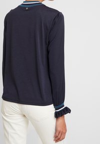 Maglietta a maniche lunghe blu navy con colletto rotondo. Presenta dettagli a righe sul colletto e sui polsini. Il tessuto è morbido con una leggera lucentezza.