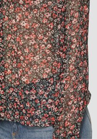 Blouse noire transparente avec un motif floral rouge et blanc, dotée de détails délicats sur le tissu et d'une coupe ample.