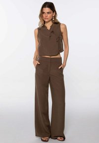 Femme portant un crop top sans manches marron avec des liens et un pantalon large assorti, se tenant avec les mains dans les poches sur fond blanc.