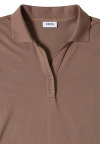 Polo marron avec col, deux boutons sur la patte de boutonnage, et une étiquette blanche avec l'inscription "CECIL" au niveau de l'encolure.