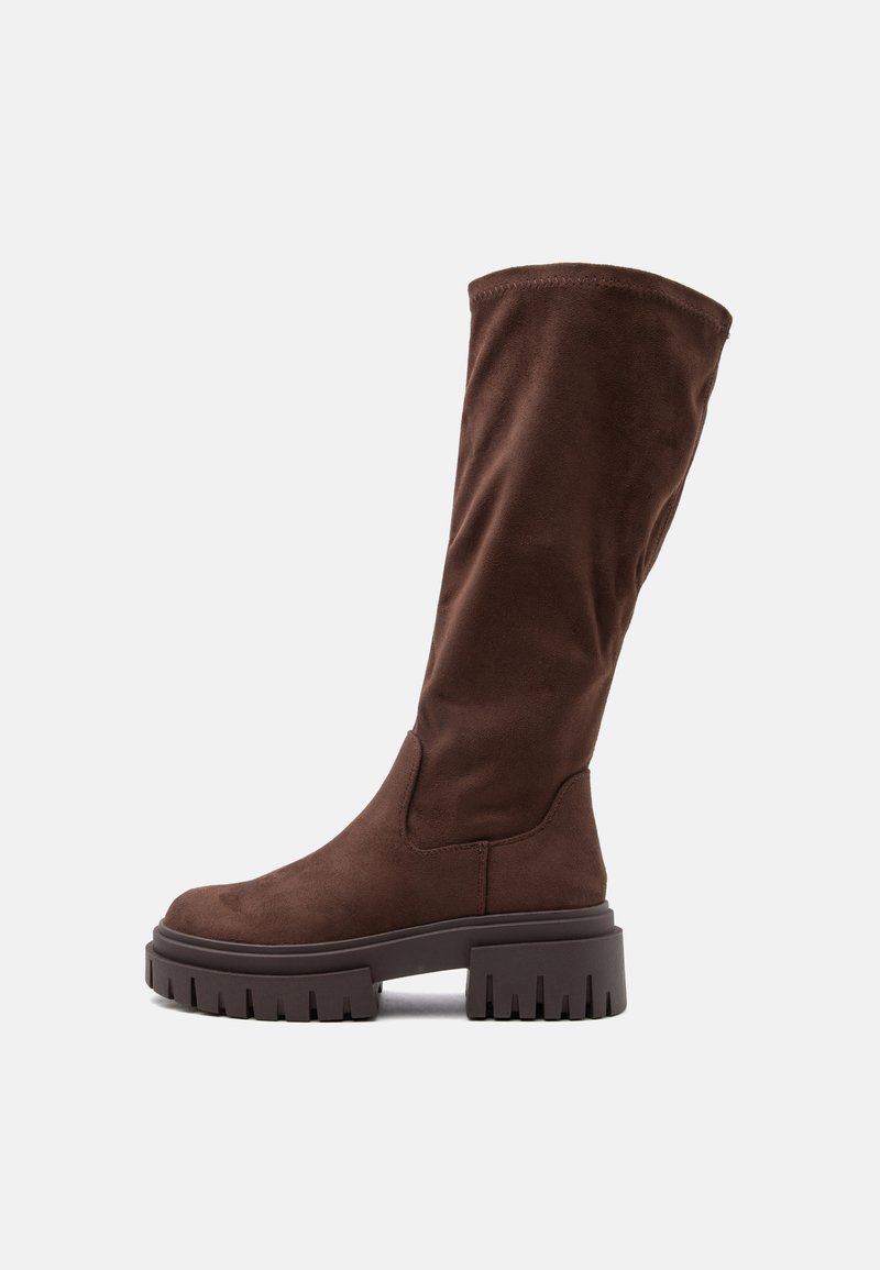 Botte haute en daim marron avec semelle épaisse à crampons et talon bas et épais vue de côté sur un fond clair uni.