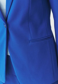 Blue blazer with textured fabric, fitted design, single pocket with subtle trim, and a button accent near the lapel.