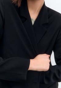 Blazer noir sur mesure avec une texture lisse, équipé d'un revers cranté et de détails subtils au niveau des coutures. Les manches sont légèrement fuselées.