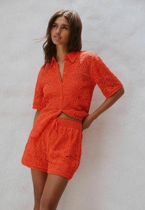 Frau trägt ein leuchtend orangefarbenes, florales Spitzenhemd mit Knöpfen und passende Shorts, posiert mit der Hand in der Hüfte vor einem hellen Wandhintergrund.