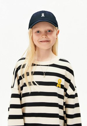 Jeune fille aux longs cheveux blonds, portant une casquette noire et une chemise rayée noir et blanc avec un patch jaune, souriante sur fond blanc.