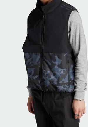 Person trägt eine schwarze und blaue Camouflage-Weste mit Reißverschluss über einem grauen Langarmshirt, schwarze Hose, silbernes Armband und Ringe, steht mit entspannten Armen.