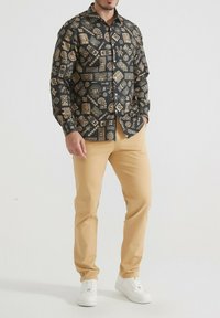 Chemise noire avec des motifs géométriques et floraux dorés, design à boutons, manches longues, assortie à un pantalon beige et des baskets blanches.