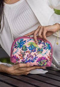 Vrouw in het wit die een roze portemonnee vasthoudt met een kleurrijk patroon van bloemen en libellen, zittend op een houten bankje in de buitenlucht.