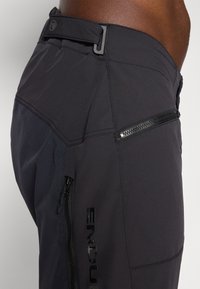 Schwarze Outdoor-Hosen mit einem eleganten Design, ausgestattet mit einer Reißverschlusstasche und einem reflektierenden Logo, aus strapazierfähigem, strukturiertem Stoff gefertigt.