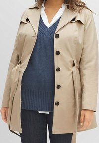 Beige trenssitakki, jossa on vyötäröllä vy�ö, suuret napit ja klassinen kaulus, päällä tummansininen neulepusero ja valkoinen paita.