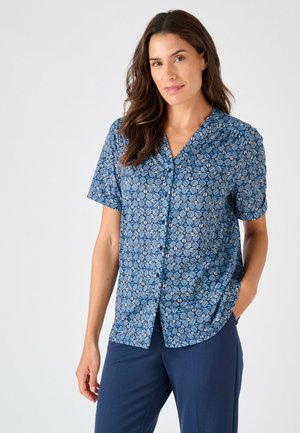 Femme aux longs cheveux bruns porte une chemise bleue à manches courtes à motifs avec boutons et un pantalon bleu marine, debout avec une main dans la poche sur un fond uni.