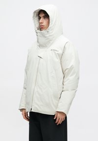 Isik, kes kannab heledat beeži kapuutsiga õueriiet, millel on tõmblukk ja TERREX logo, koos mustade pükstega, seistes ühtlase tausta ees.