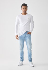 Camisa blanca de manga larga con un pequeño logo amarillo, combinada con jeans de lavado claro y zapatillas blancas. Ajuste moderno con una textura de tela suave.