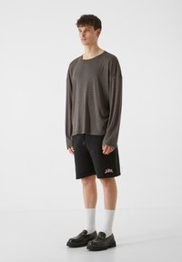 Dlhý rukáv, tmavošedá košeľa s voľným strihom, doplnená o čierne šortky s ružovým logom. Model nosí biele ponožky a čierne topánky.