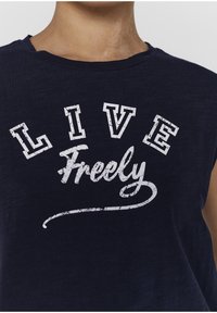 Námořnická modrá bezrukávová bavlněná trička se silným, opotřebovaným bílým nápisem: "ŽIJ" nad kurzívou "Svobodně". Hladká textura.